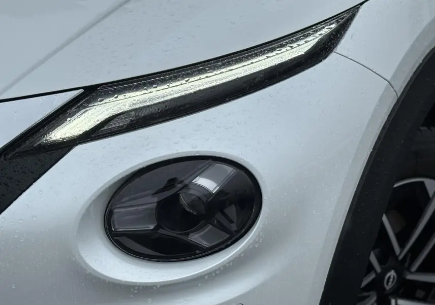 Gros plan sur le phare avant droit et la jante alliage du Nissan Juke blanc nacré sous la pluie.