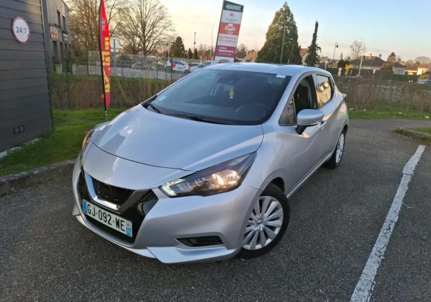 Nissan Micra gris clair vue 3/4 avant droit avec phares allumés, stationnée sur un parking extérieur.