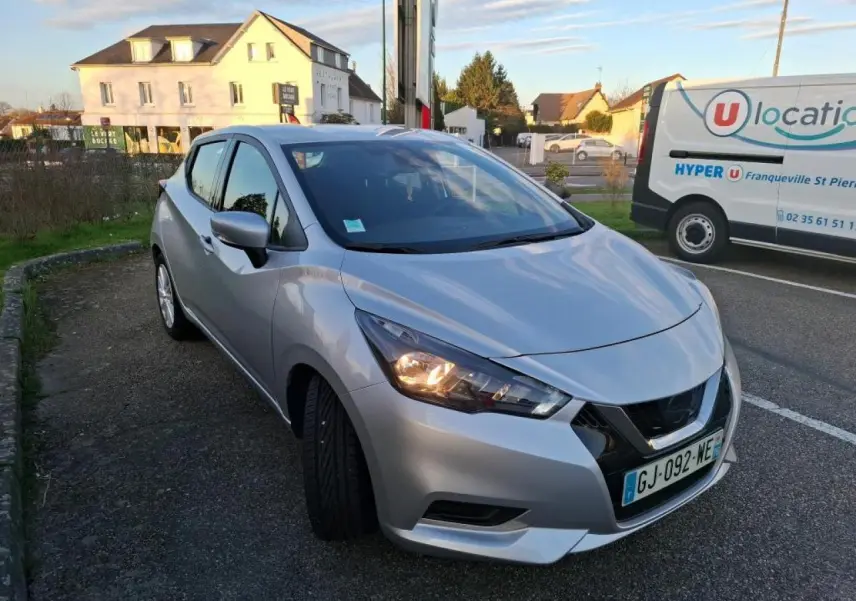Nissan Micra gris clair vue 3/4 avant droit, phare avant allumé, stationnée sur un parking extérieur.