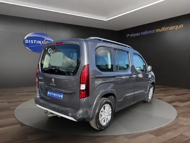 Vue 3/4 arrière droite d’un Peugeot Rifter gris foncé stationné en intérieur avec barres de toit chromées visibles.