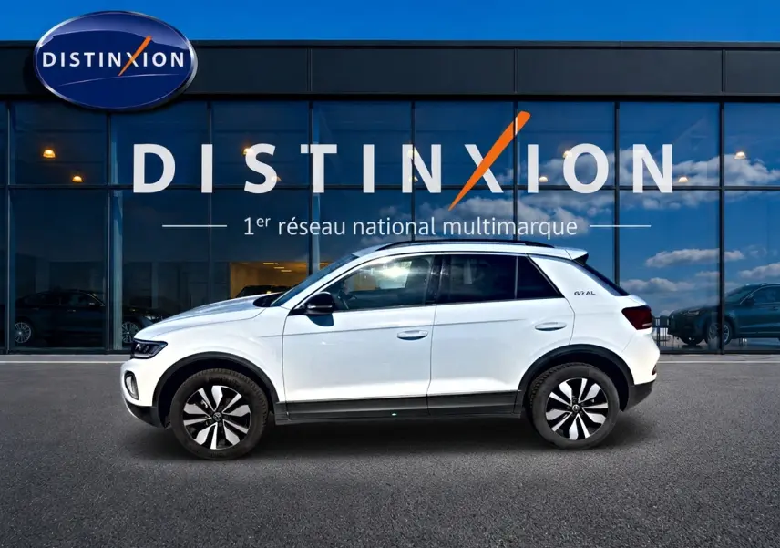 Volkswagen T-Roc blanc en profil droit, avec jantes alliage 17 pouces et barres de toit visibles, devant une vitrine Distinxion.