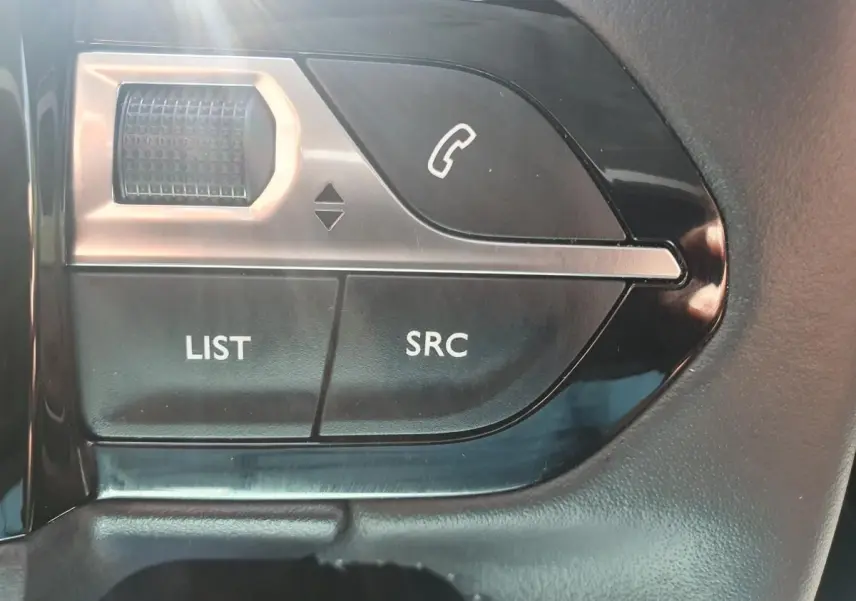 Gros plan sur les commandes au volant côté gauche de la Peugeot 208 gris Artense, boutons LIST, SRC et téléphone.