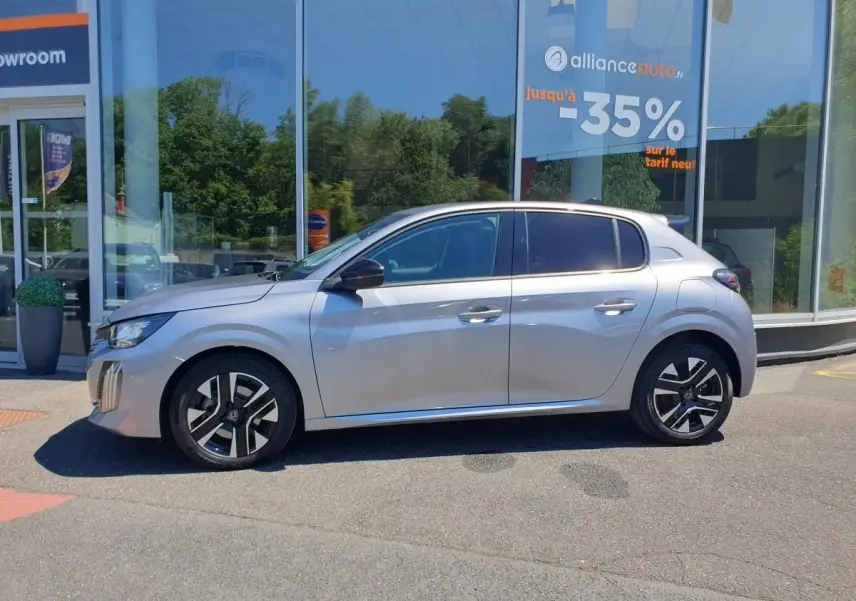Profil droit de la Peugeot 208 1.2 Hybrid 110 Allure gris Artense avec jantes alliage 16'' bi-tons devant un showroom.