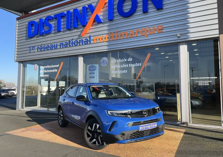 Opel Mokka bleu Voltak en 3/4 avant droit, devant une concession Distinxion avec calandre noire et jantes stylées.