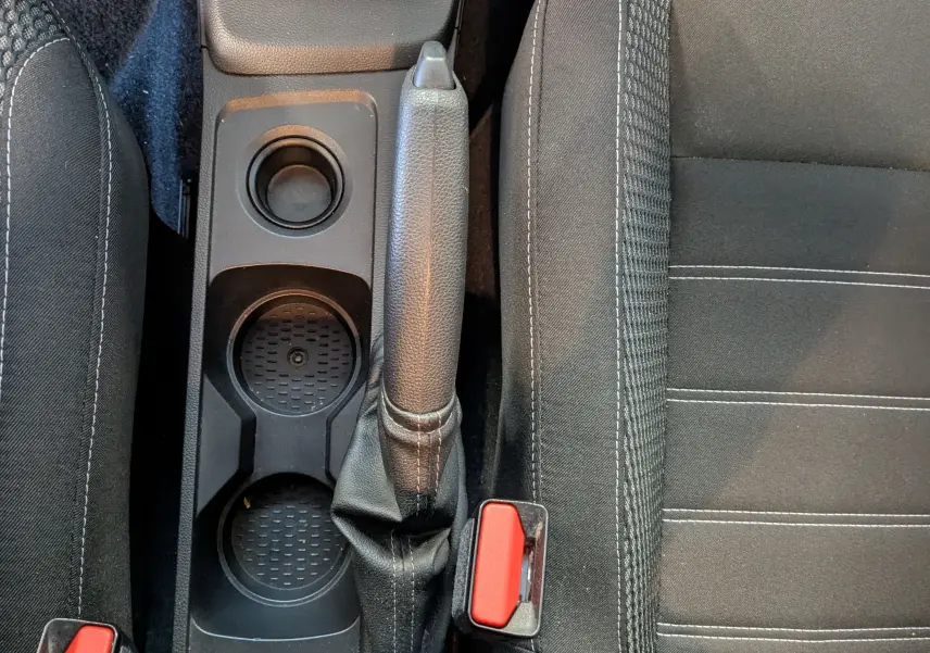 Vue plongeante sur la console centrale du Dacia Duster 2023, montrant frein à main et porte-gobelets noirs entre sièges tissu noir.