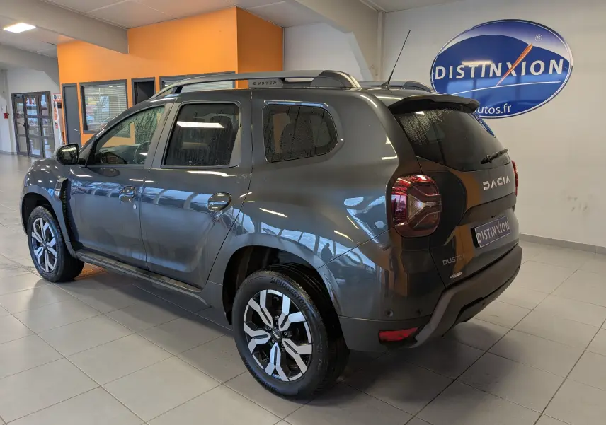Vue 3/4 arrière droite d'un Dacia Duster gris 2023 en intérieur, avec jantes bi-ton et barres de toit visibles.