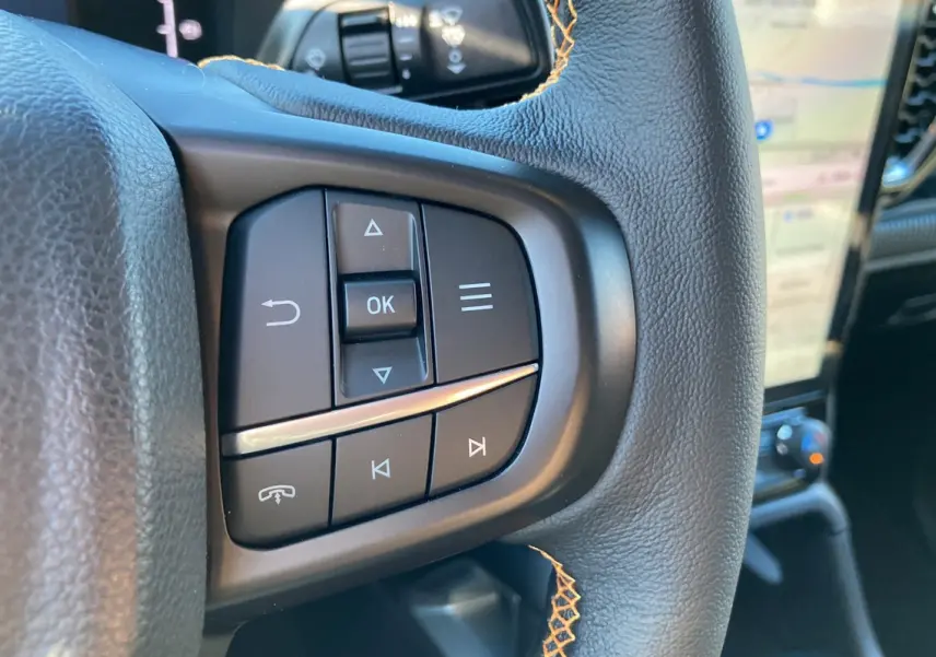 Gros plan sur les commandes multifonctions en cuir noir du volant du Ford Ranger Wildtrak blanc glacier, avec écran tactile en arrière-plan.