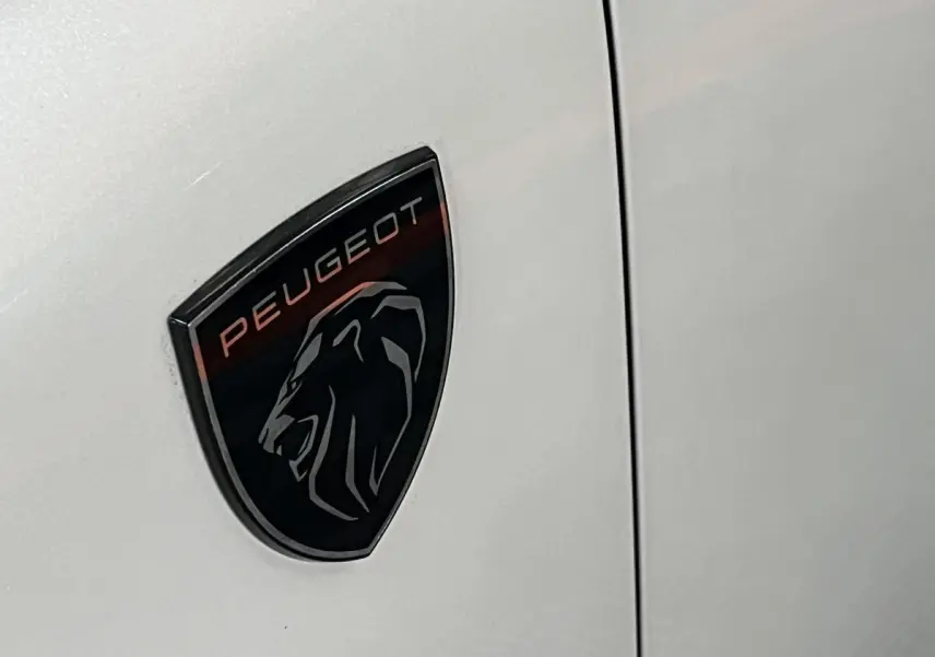 Gros plan sur l'écusson Peugeot noir et rouge sur la carrosserie blanc nacré d'une 308 GT PHEV.