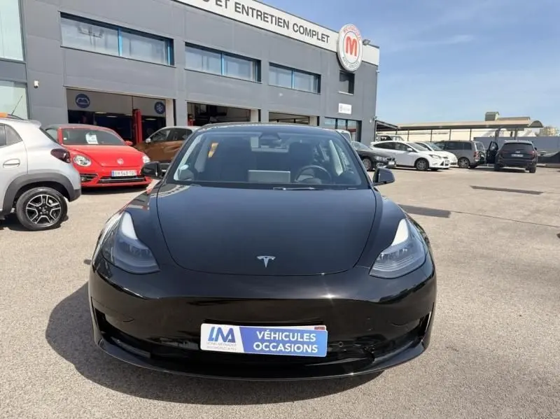 Vue avant d'une Tesla Model 3 Standard RWD Plus noire 2022, garée devant un garage avec plaque "Véhicules Occasions".