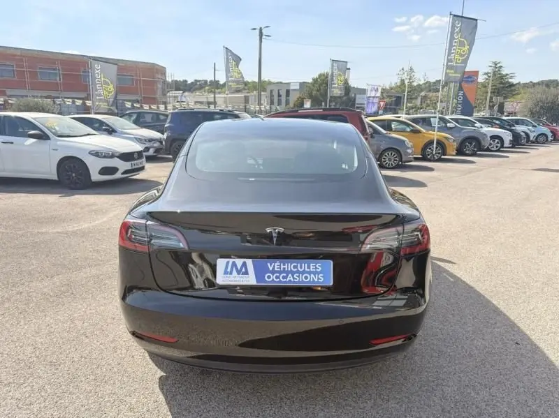 Vue arrière d'une Tesla Model 3 Standard RWD Plus noire 2022, stationnée en extérieur sur un parking de véhicules d'occasion.