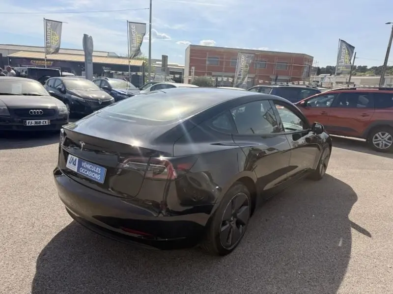 Tesla Model 3 Standard RWD Plus noire vue en 3/4 arrière droit sur un parking ensoleillé, avec jantes noires spécifiques.