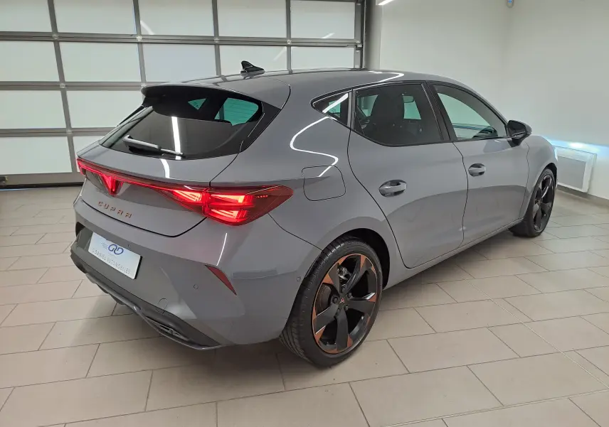 Vue 3/4 arrière droite d'une CUPRA Leon 2025 gris Graphène avec jantes cuivrées et toit ouvrant panoramique en intérieur.