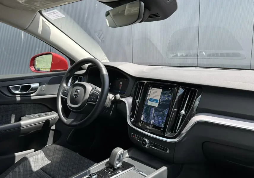 Vue intérieure côté conducteur de la Volvo S60 rouge Fusion 2024, avec tableau de bord et écran tactile GPS visible.