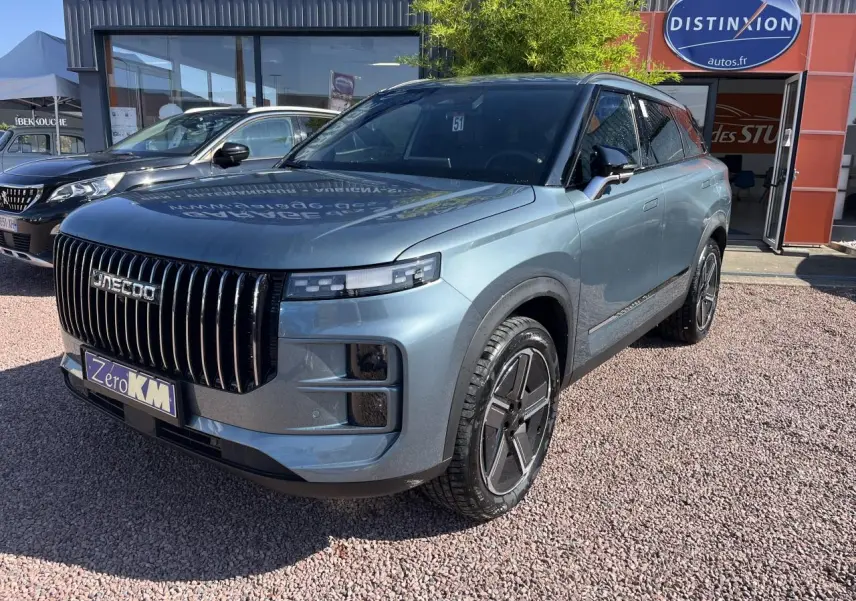 Vue 3/4 avant d'un JAECOO J7 gris olive avec calandre verticale noire et jantes modernes, stationné devant un showroom.