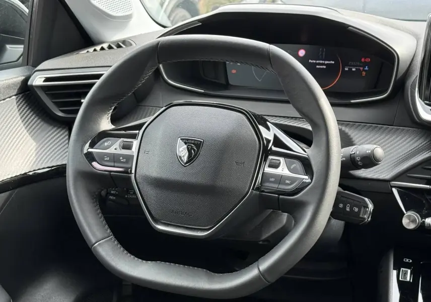 Vue rapprochée du volant noir de la Peugeot 208 2025 avec tableau de bord digital et détails carbone autour.