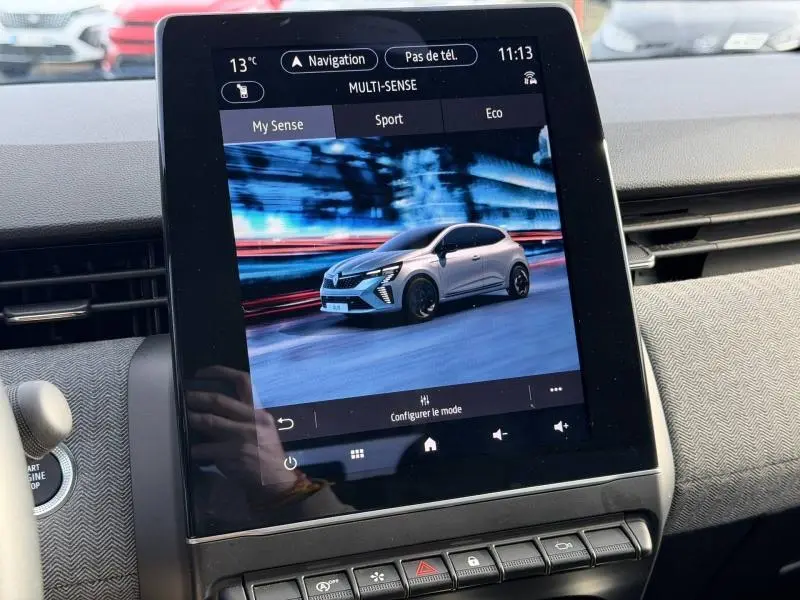 Écran tactile intérieur montrant une Renault Clio blanche en 3/4 avant sur fond urbain flouté.