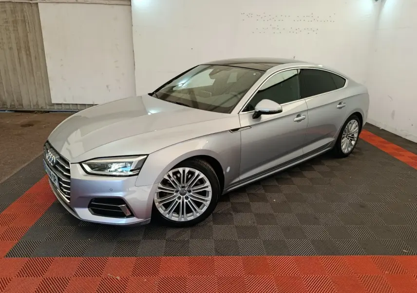 Audi A5 gris clair vue 3/4 avant droit dans un garage avec sol noir et rouge, jantes alliage multibranches.