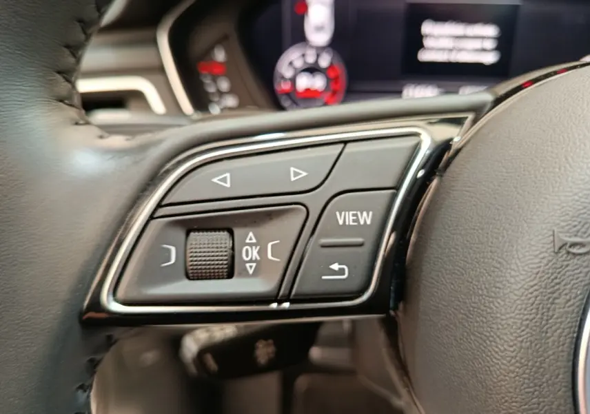 Gros plan sur les commandes multifonctions côté gauche du volant d’un Audi A5 gris clair, tableau de bord flou en arrière-plan.
