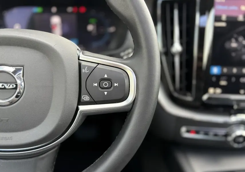 Gros plan sur le volant noir en simili-cuir du Volvo XC60 2024 avec commandes intégrées et tableau de bord flou en arrière-plan.