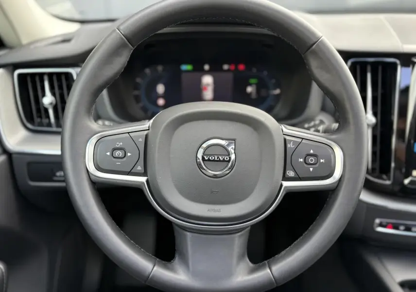Vue rapprochée du volant simili-cuir noir du Volvo XC60 2024 avec commandes intégrées et tableau de bord digital flou.