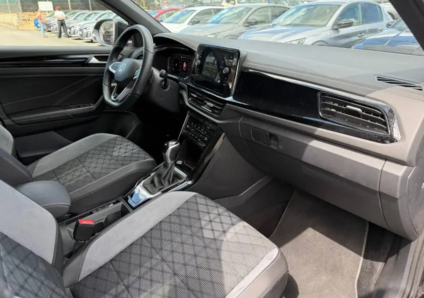 Vue intérieure côté conducteur du Volkswagen T-Roc 1.5 TSI R-Line 2025, sièges tissu gris et tableau de bord noir brillant.