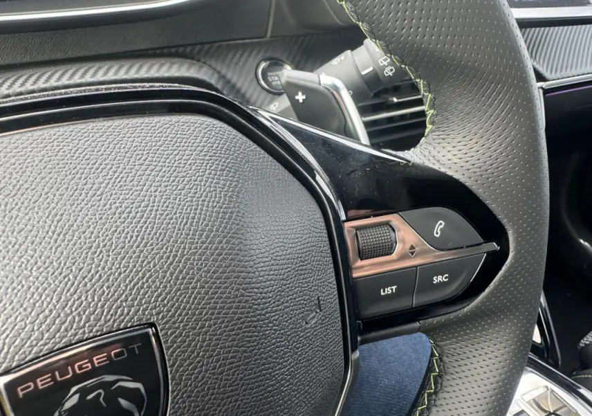 Gros plan sur le volant perforé avec surpiqûres vertes du Peugeot 208 Hybrid 110 e-DCS6 GT, montrant les commandes audio et téléphone.
