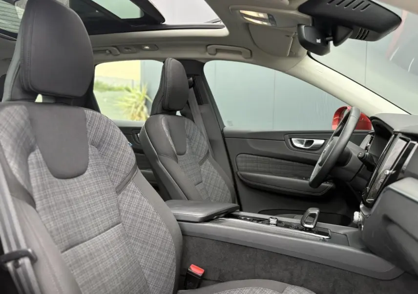 Vue intérieure côté conducteur du Volvo XC60 2024, sièges tissu gris à motif, toit ouvrant panoramique et volant simili-cuir.