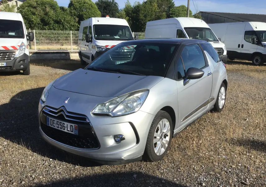Citroën DS3 gris clair métal vue 3/4 avant droit sur un parking avec plusieurs fourgons blancs en arrière-plan.