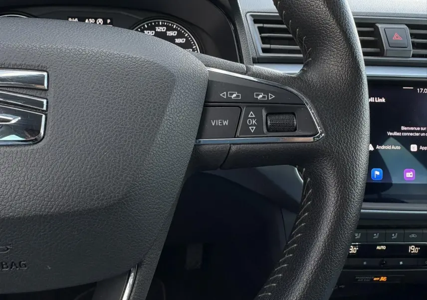 Gros plan sur le volant en cuir noir du SEAT Arona gris magnétique, avec commandes multifonctions et écran tactile en arrière-plan.