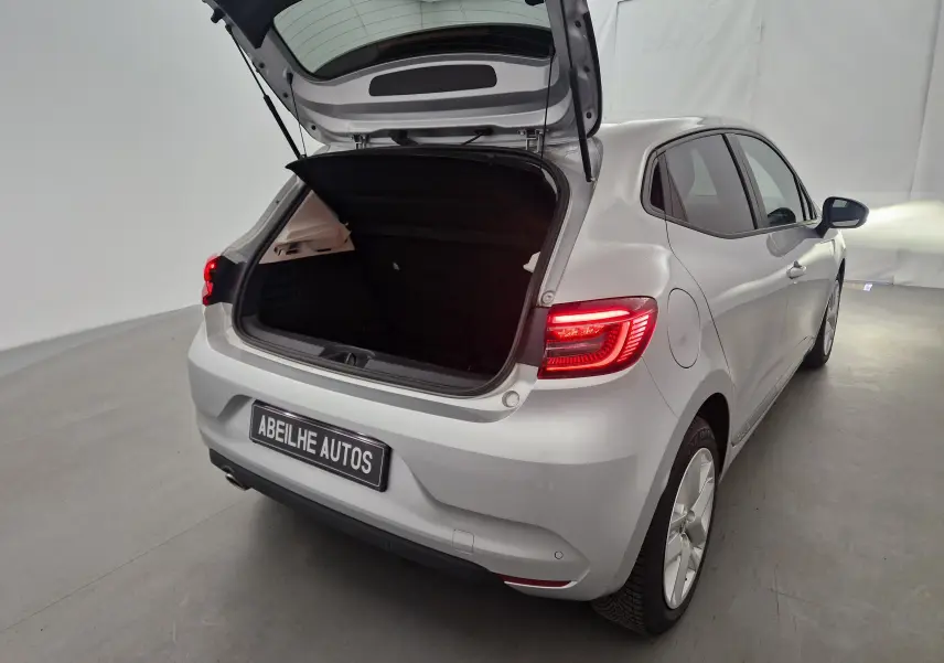 Renault Clio V gris platine vue 3/4 arrière droit avec coffre ouvert et feux arrière allumés dans un studio.