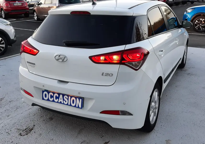 Vue 3/4 arrière droite d'une Hyundai i20 blanche de 2016 avec feux allumés et plaque "OCCASION" visible.