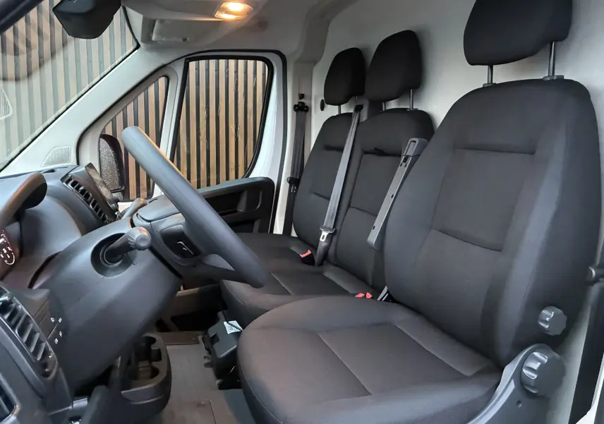 Vue intérieure côté conducteur du Fiat Ducato Fourgon 2.2 blanc, montrant les sièges noirs en tissu et le tableau de bord.