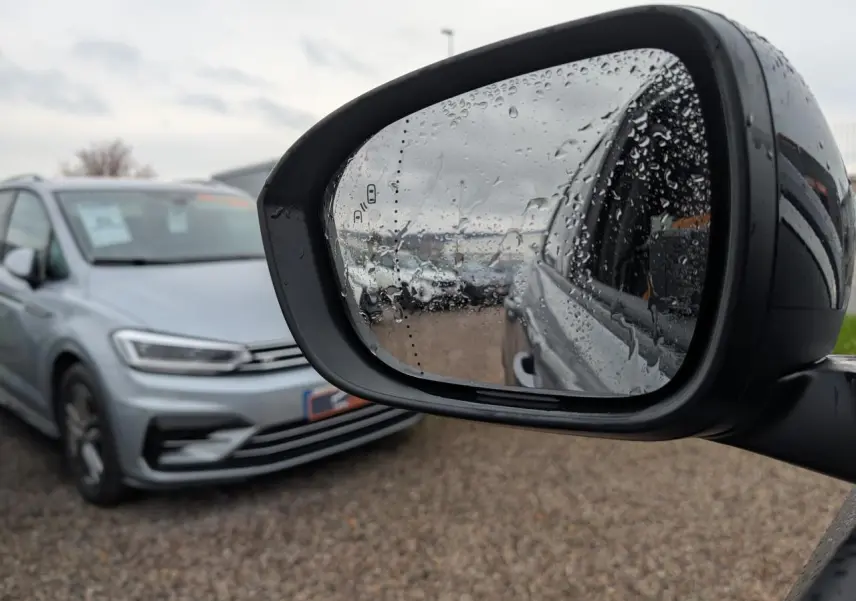 Rétroviseur côté droit avec gouttes d'eau reflétant un véhicule gris en stationnement, vue extérieure floue.