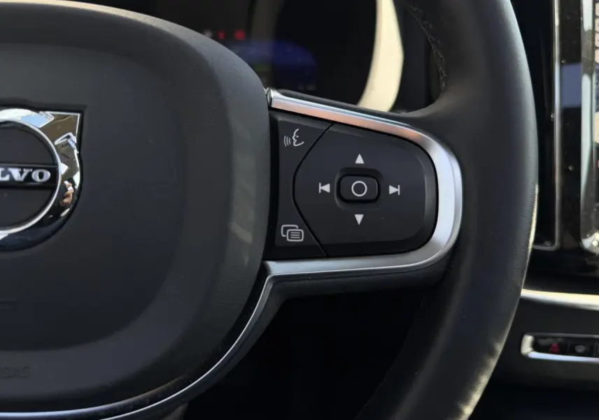 Gros plan sur le côté droit du volant noir simili-cuir du Volvo XC60 2024 avec commandes intégrées et logo chromé.