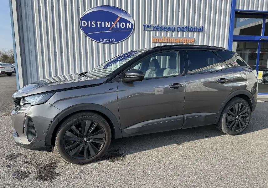 Peugeot 3008 gris en profil droit, finition GT avec jantes noires et toit noir, stationné devant un bâtiment commercial.