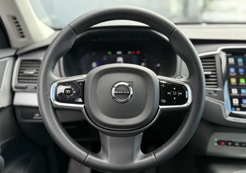 Vue rapprochée du volant noir du Volvo XC90 T8 2024 avec commandes intégrées et tableau de bord digital flou en arrière-plan.