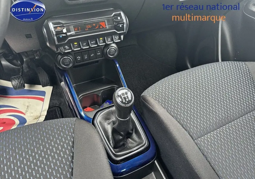Vue intérieure centrée sur la console centrale et levier de vitesse manuelle, sièges tissu gris avec motifs bleus.