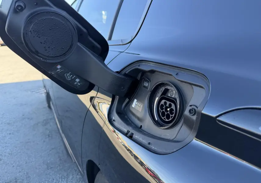 Prise de recharge ouverte côté gauche d’une Peugeot 308 noire métallisée, avec reflets sur la carrosserie.