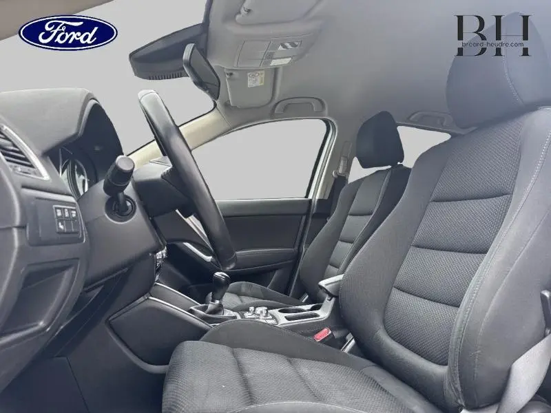 Intérieur du Mazda CX-5 2016 vu côté conducteur, sièges noirs et tableau de bord avec volant et levier de vitesse manuel.