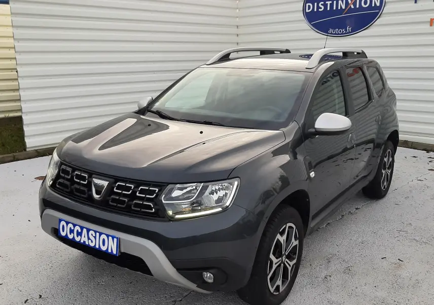 Dacia Duster gris Cassiopée en 3/4 avant droit, avec jantes bi-ton et barres de toit visibles.
