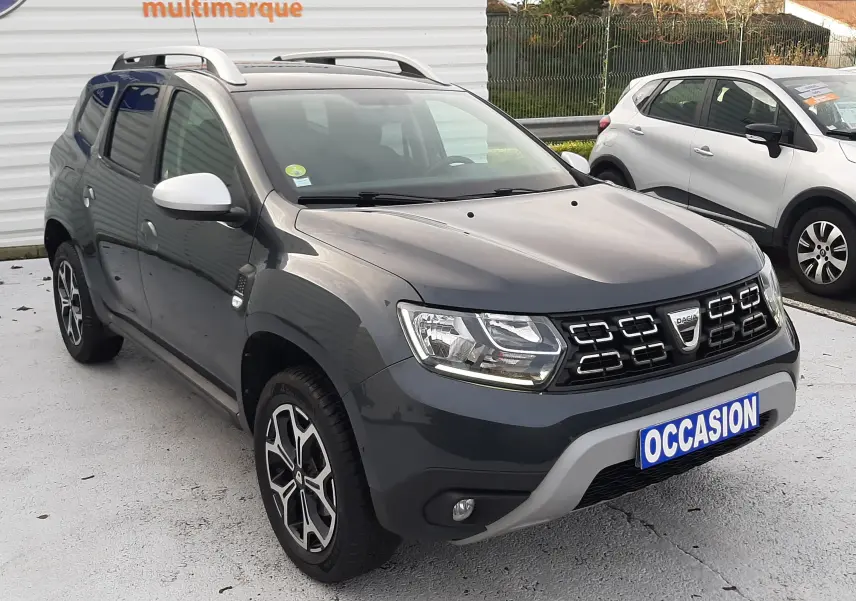 Dacia Duster gris cassiopée en 3/4 avant droit, avec jantes bi-ton et barres de toit visibles sur parking.