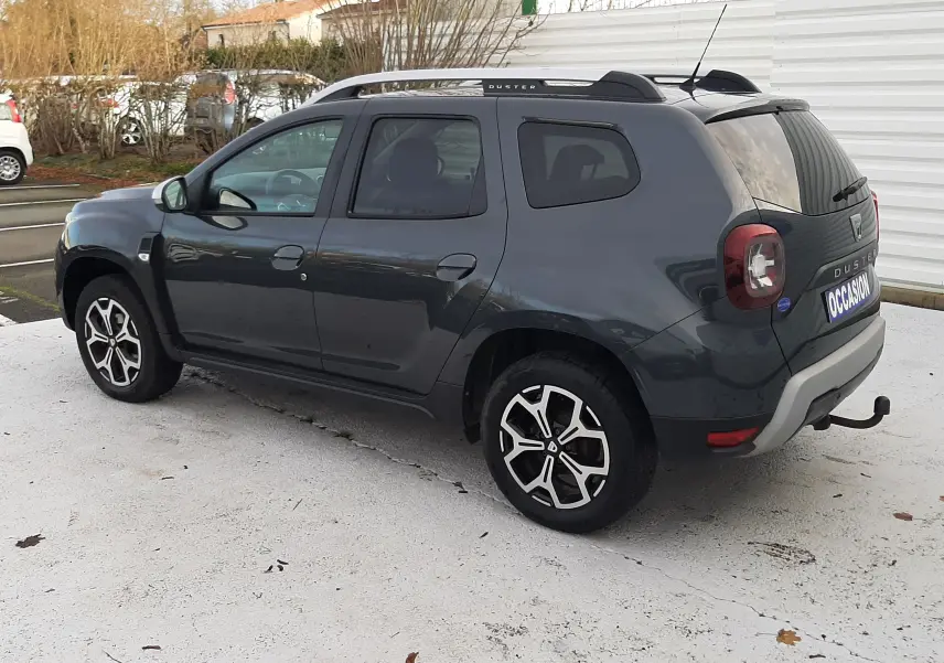 Vue 3/4 arrière droite d'un Dacia Duster gris Cassiopée avec jantes bi-ton et attelage visible sur parking extérieur.