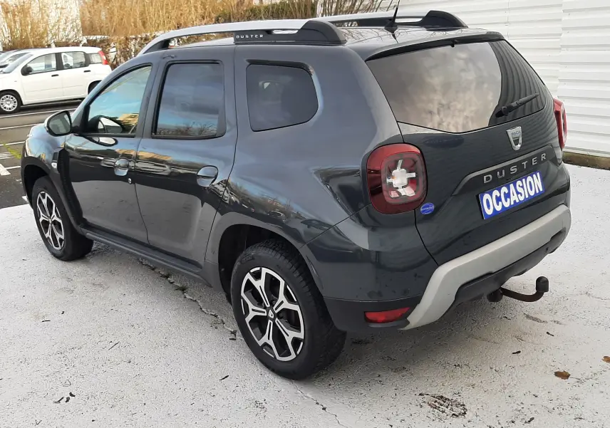 Vue 3/4 arrière droite d'un Dacia Duster gris Cassiopée avec barres de toit noires et attelage visible.