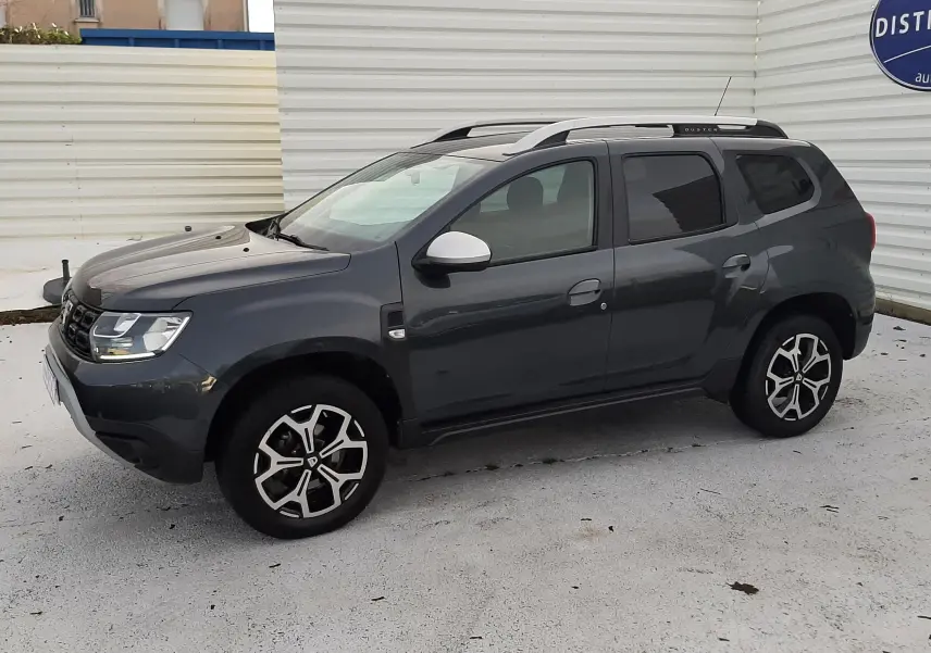 Vue latérale gauche d'un Dacia Duster gris Cassiopée 2019 avec jantes alliage bicolores et barres de toit.