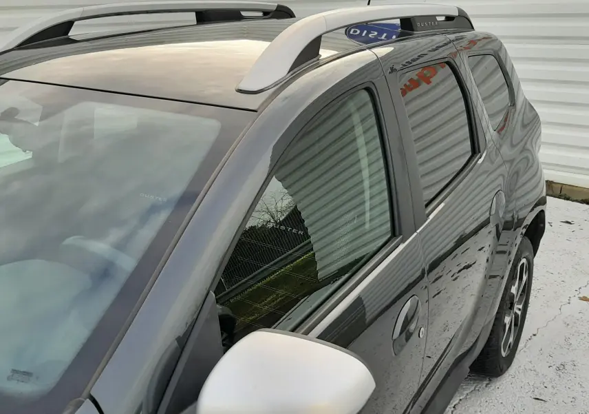 Vue 3/4 avant droit du Dacia Duster gris Cassiopée, avec barres de toit blanches et rétroviseur blanc contrastant.