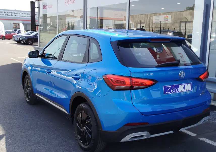 Vue 3/4 arrière gauche du SUV MG ZS Cosmo Blue 2026 avec jantes noires et feux arrière LED, stationné devant un showroom.