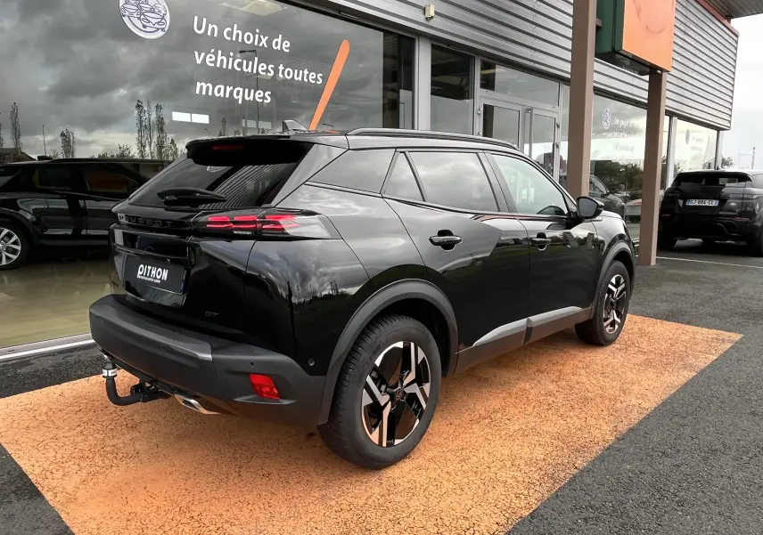 Peugeot 2008 noir en 3/4 arrière droit, avec jantes distinctives et attelage visible, stationné devant un showroom.
