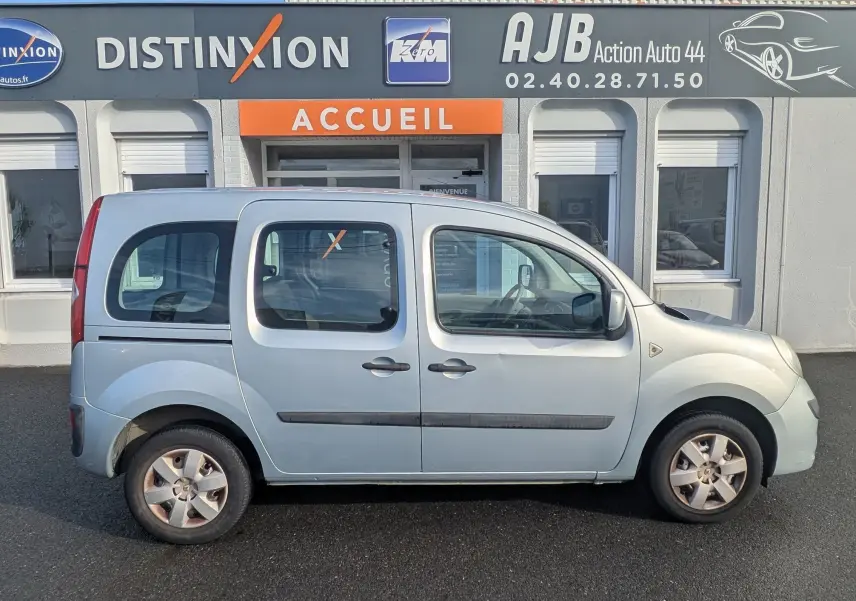 Profil latéral droit d'un Renault Kangoo II bleu ciel stationné devant un bâtiment commercial avec portes coulissantes visibles.