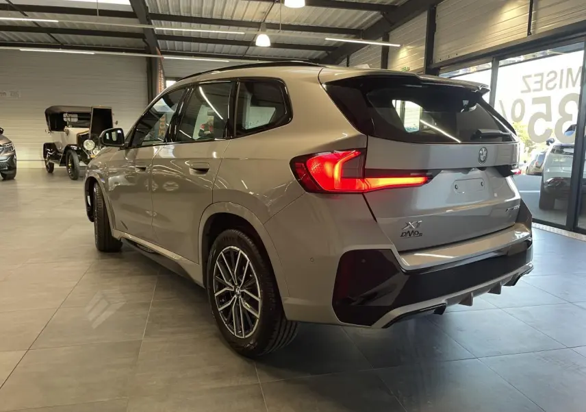 BMW X1 xDrive 20d 2024 en Spacesilber Metallic, vue 3/4 arrière droite avec feux LED allumés et jantes M Sport.