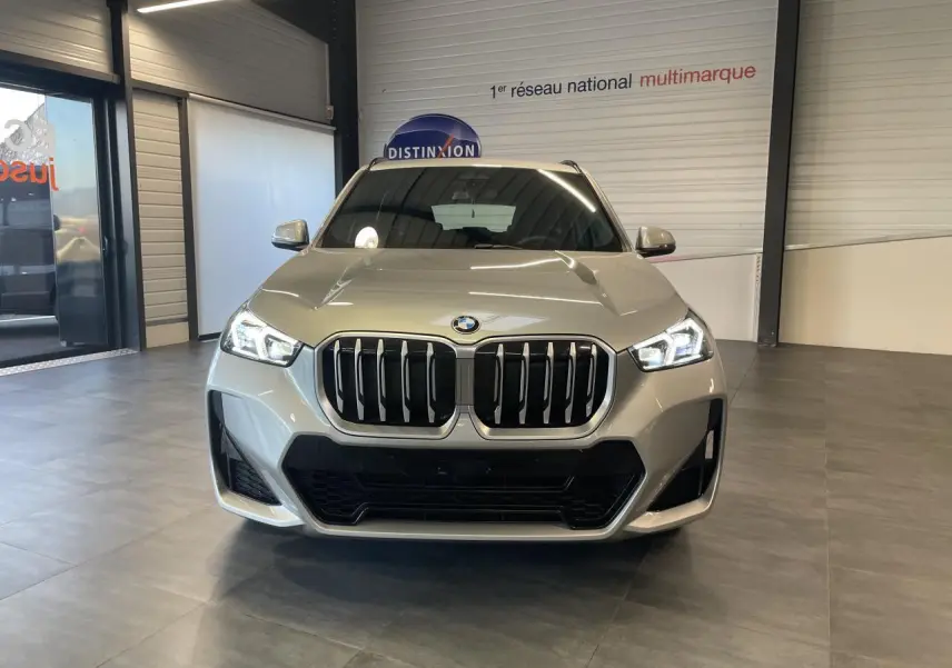 Vue frontale d'un BMW X1 xDrive 20d 2024 en couleur Spacesilber Metallic avec calandre M Sport et phares LED allumés.